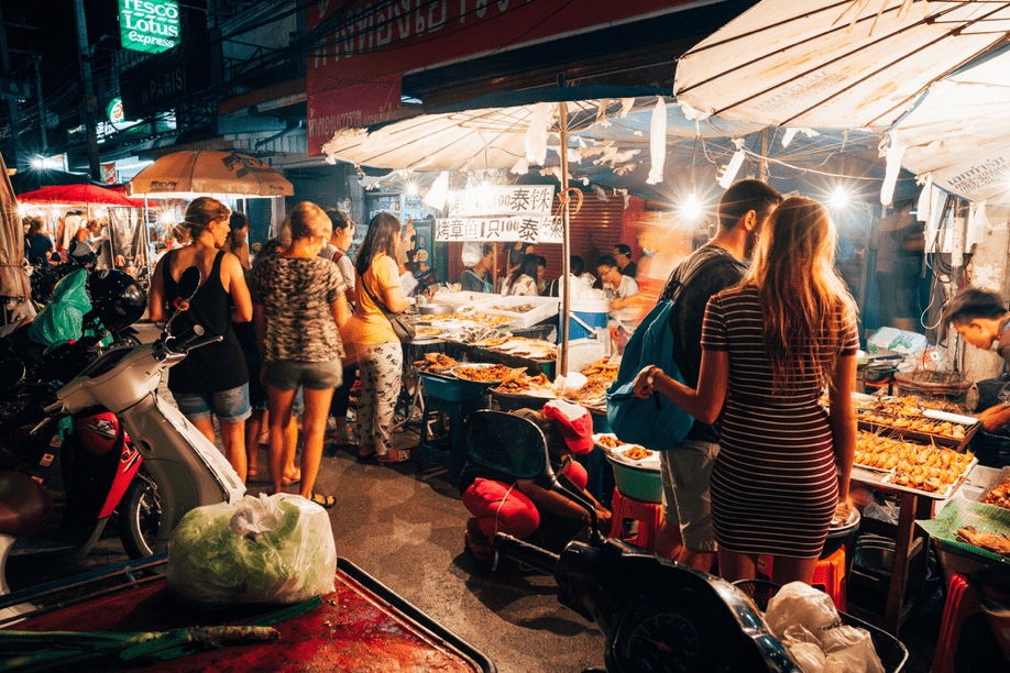 People shopping in night market filled with food. Motorbikes on the left side, several food stalls on the right with shoppers browsing.