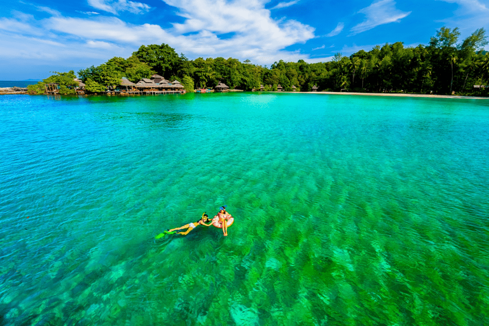 People snorkeling and sitting in inner tube floating above emerald water. Beach covered with sand next to trees, and small villas are in the background.