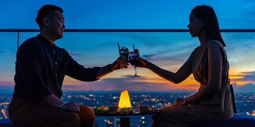 Couple cheering with drinks on a rooftop bar in Bangkok during sunset